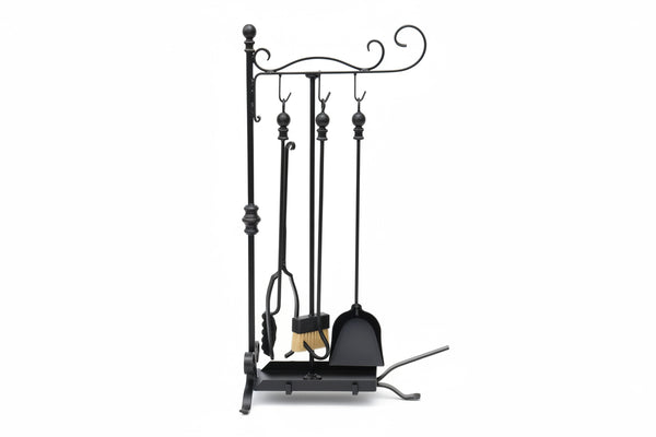 Black wrought iron fireplace tool set with a poker, tongs, brush, and stand, photographed outdoors against a concrete pillar on patterned tile flooring.