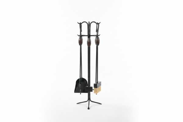 A black wrought-iron fireplace tool set with wooden handles and a light tan brush bristle head, displayed on a stand in front of a large concrete pillar.